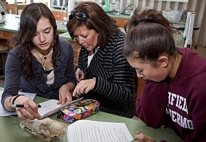 Image shows a woman with her two daughters on either side of her, reading an essay that one of them is presenting to her.