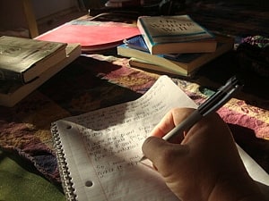 Image shows someone writing in a notebook, surrounded by books. 