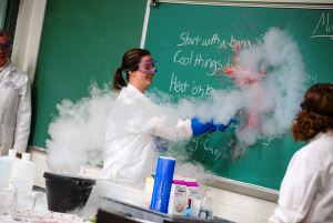 Image shows a chemistry experiment belching smoke, while a woman in a lab coat writes on a chalkboard behind it.