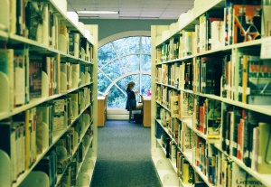 Image shows a student in a library, seen between bookshelves.